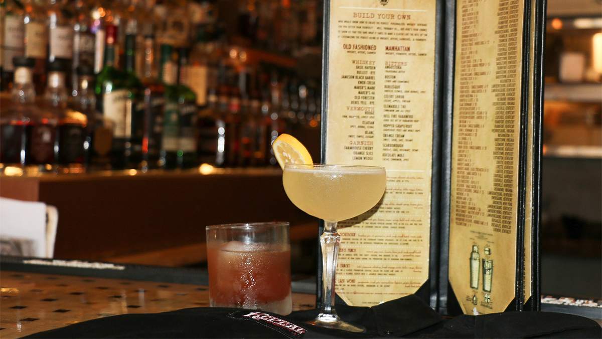 Close up of two alcoholic drinks with a tall drink menu behind them and a blurry bar in the back ground at Repeal Bourbon and Burgers in Virginia Beach, Virginia, USA